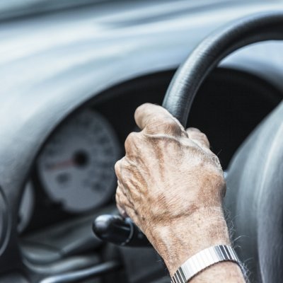 A older person with their hand on a car steering wheel.